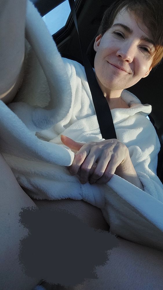 Eine Person sitzt im Auto, trägt einen weißen Bademantel und ist mit einem Sicherheitsgurt angeschnallt. Mit einer Hand wird der Mantel geöffnet, während die andere das Handy hält. Das Gesicht ist im Sonnenlicht zu sehen, mit einem leichten Lächeln. Der Blickwinkel ist von unten aufgenommen.