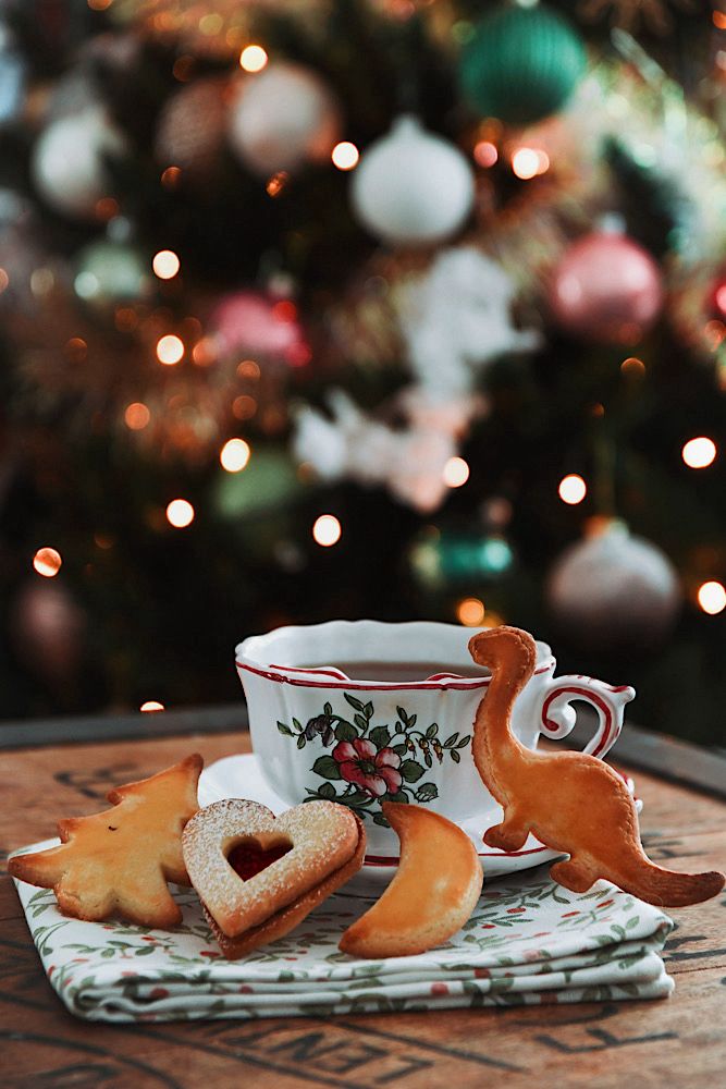 Au dernier plan un sapin de Noël décoré. Sur la table sont posés sur une serviette une tasse de thé et 4 biscuits de Noël en formes de sapin, de cœur, de lune et de dinosaure.
