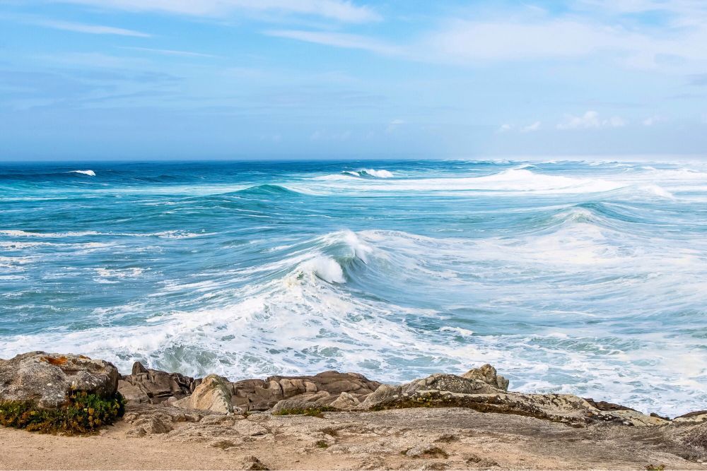 Photo format paysage prise à la pointe de la Torche dans le Finistère. Au premier plan, le promontoire rocheux de la pointe. La plage est hors-cadre au loin à droite. Les vagues déferlent de la gauche de la photo vers la droite en direction de la plage. Elles ont des nuances bleues et vertes et sont pleines d’écume.