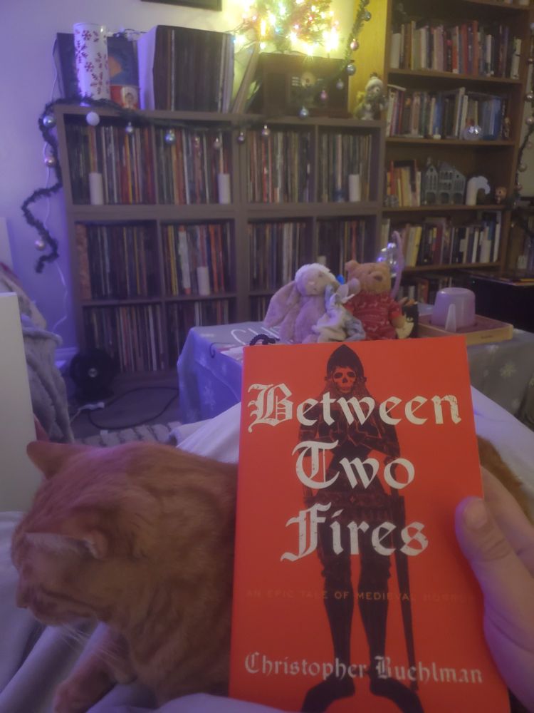 A copy of "Between Two Fires" by Christopher  Buehlman" held on my lap, leaned against a loafing orange cat, with shelves of records and books in the background. 