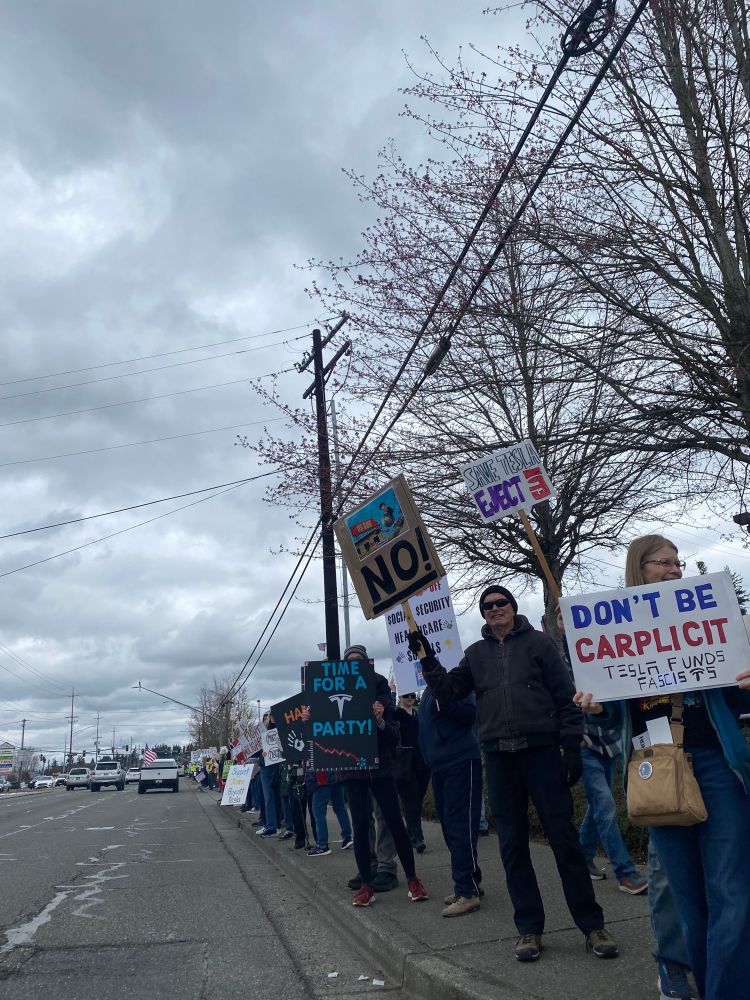 Protesters on roadside in front of Tesla dealership with sign, saying No!, don’t be carplicit, save Tesla eject Elon Tesla funds fascists 