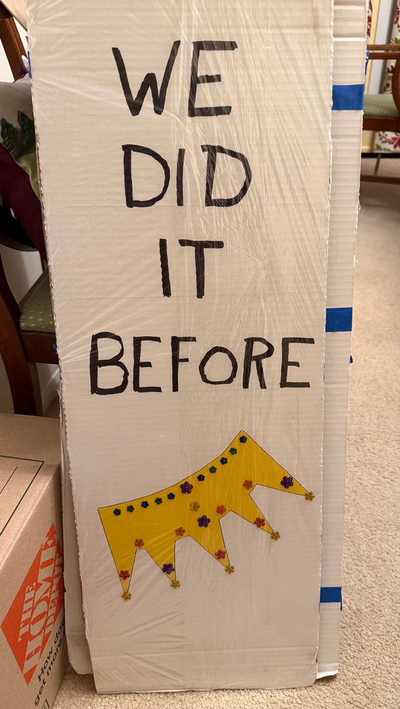 Narrow sign that says, “We did it before” with an upside down crown under the text. 