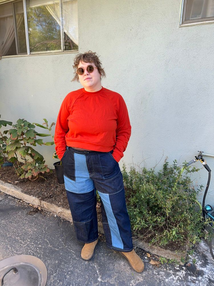 A white woman with strawberry blonde hair is standing on a patio with some foliage in the background. She’s wearing pink rimmed sunglasses, a bright tomato red raglan sweater and patchwork jeans made of different colors of denim and brown boots. Her hands are in her pockets. Sewing deets - I changed front pocket to an angled patch pocket. I lengthened the pants a few inches so they aren’t as cropped as drafted.