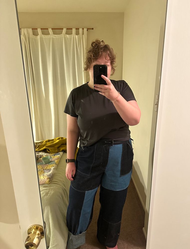 A white person is taking a selfie in a mirror with the phone covering their face. They are wearing a gray little tshirt and a pair of patchwork jeans in varying colors of denim. One of the legs is cuffed. 