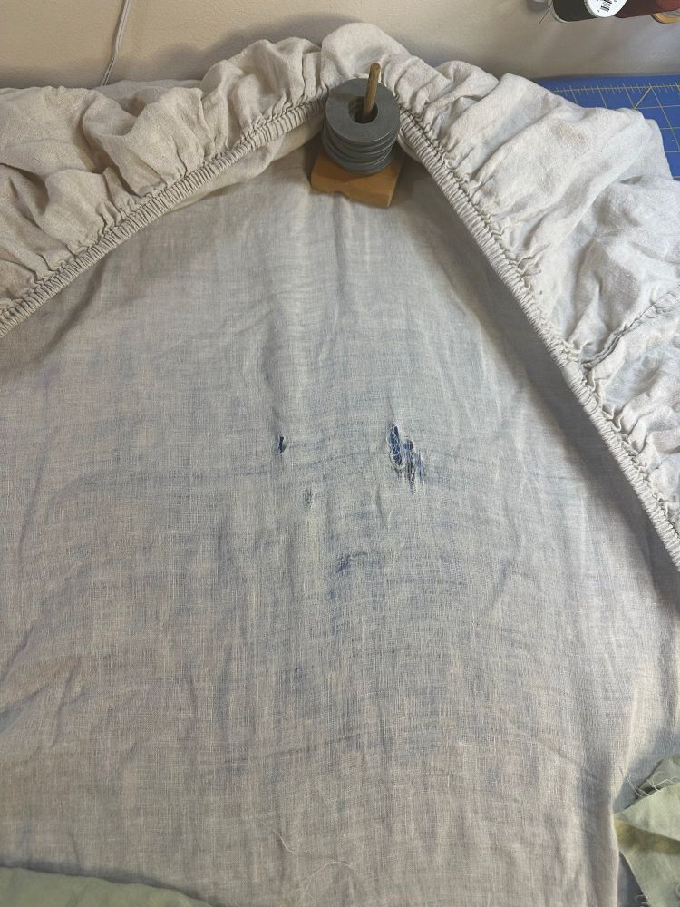 An off white linen fitted sheet has worn thin and has some holes in it. 