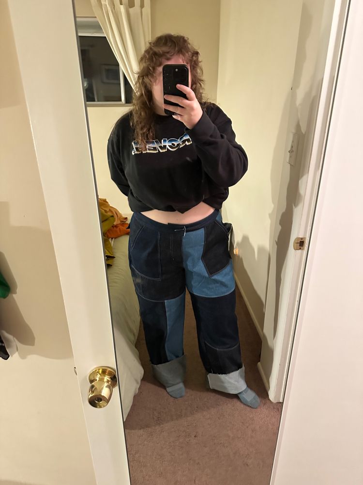 A white person with long curly hair is wearing a black sweatshirt and a pair of denim patchwork jeans they made. They are taking a picture in a mirror and their phone is covering their face. 
