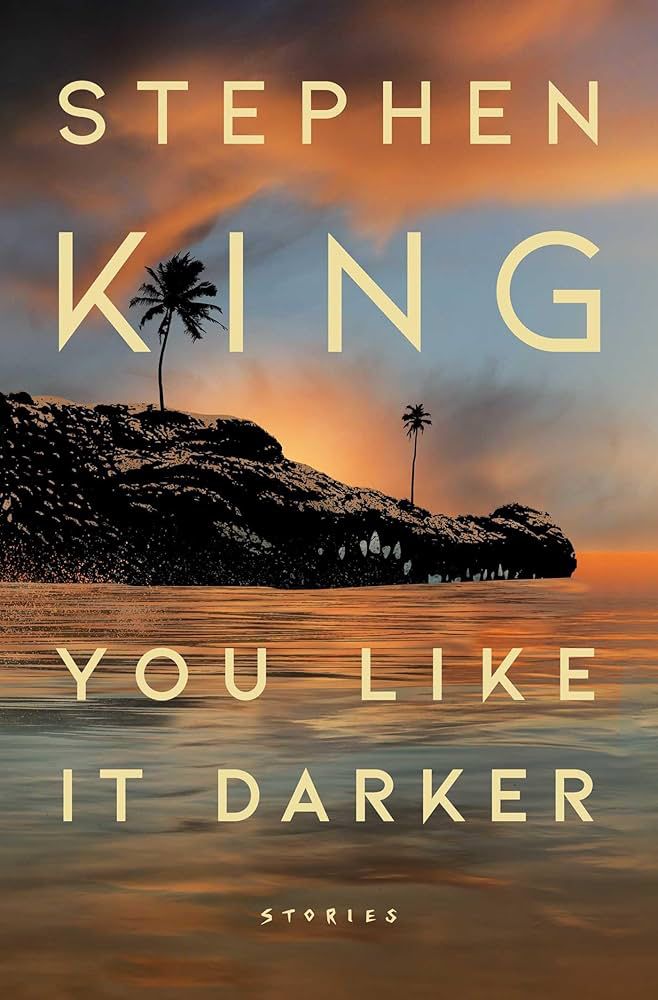 The book cover for “you like it darker” by Stephen king. The background is of a sunset over a body of water and a crocodile (alligator? Idk the difference) peeking out with two palm trees on its head. It’s meant to look like an island from far away. 