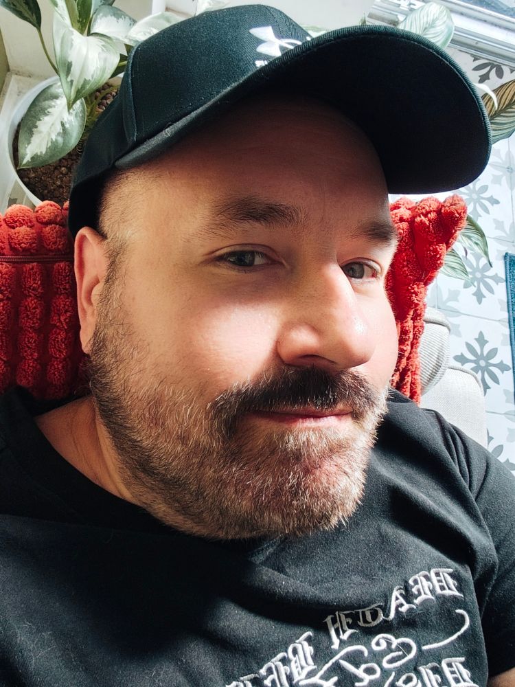Selfie. I'm a bear man with mustache and beard wearing a black shirt and a black cap. I'm resting on a red pillow. There are houseplants behind me.