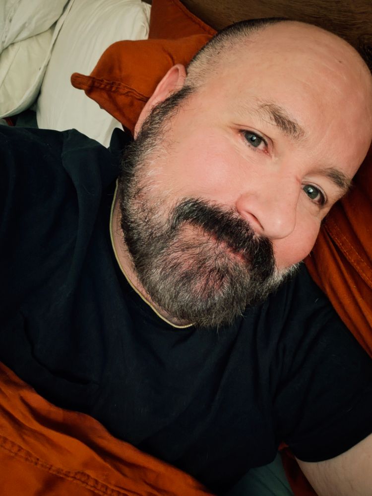 Selfie. I'm a bear man with mustache and beard wearing a black shirt and nestled in orange blankets.