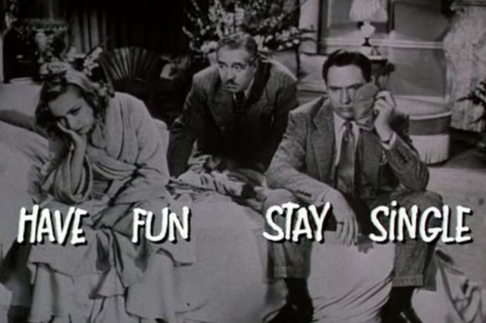 Black and white photo man in a suit on right sitting on bed with ice bag on his face, a woman in a bathrobe on opposite end of bed has her head in her hand, another man in a suit is leaning in between from behind them. The words Have Fun Stay Single in white letters across image