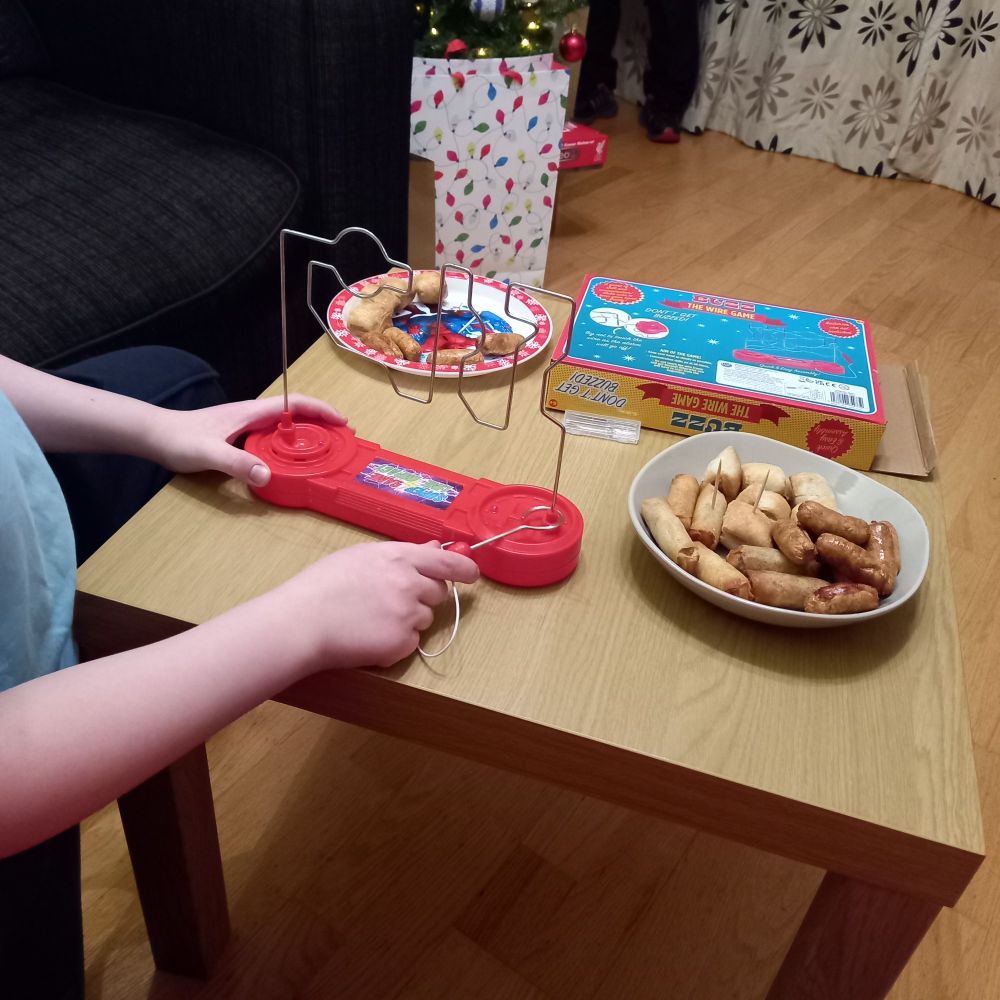 A living room. A child plays a game of Buzz the Wire Game. Next to him is a bowl of sausages and various beige air fryer foods. 