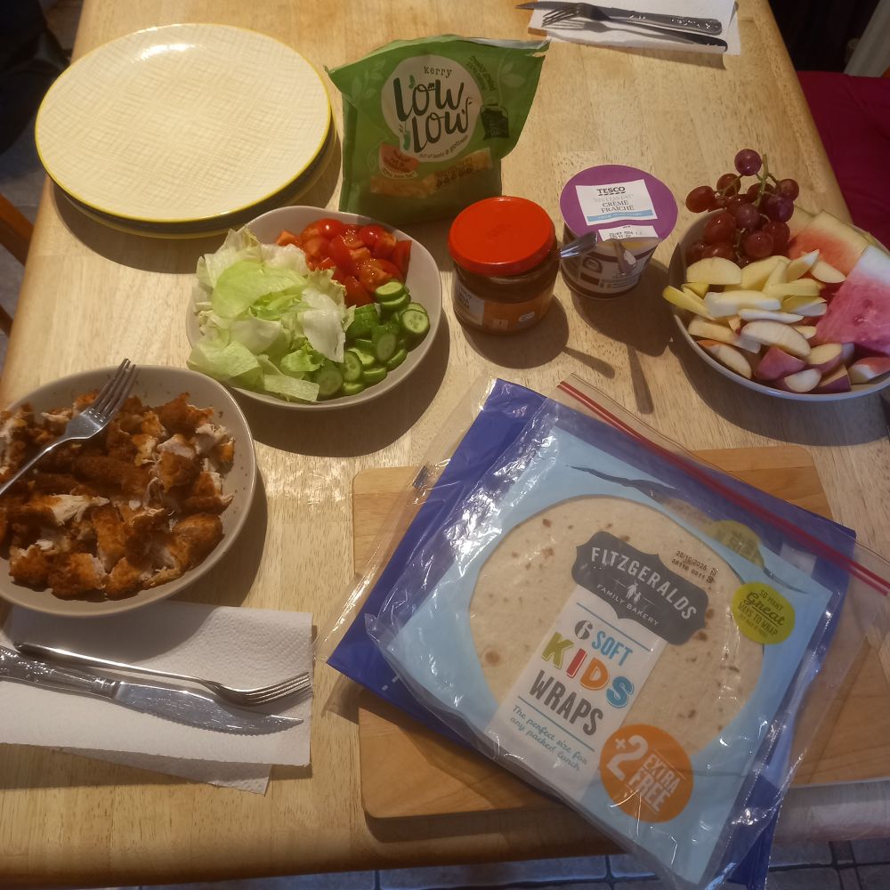 A table set with fixins for chicken wraps. In bowls we have hot breaded chicken, tomato, cucumber and lettuce. Then a bowl with melon, grapes, peach and apple. There is also cheese, sour cream, salsa and wraps on the table. 