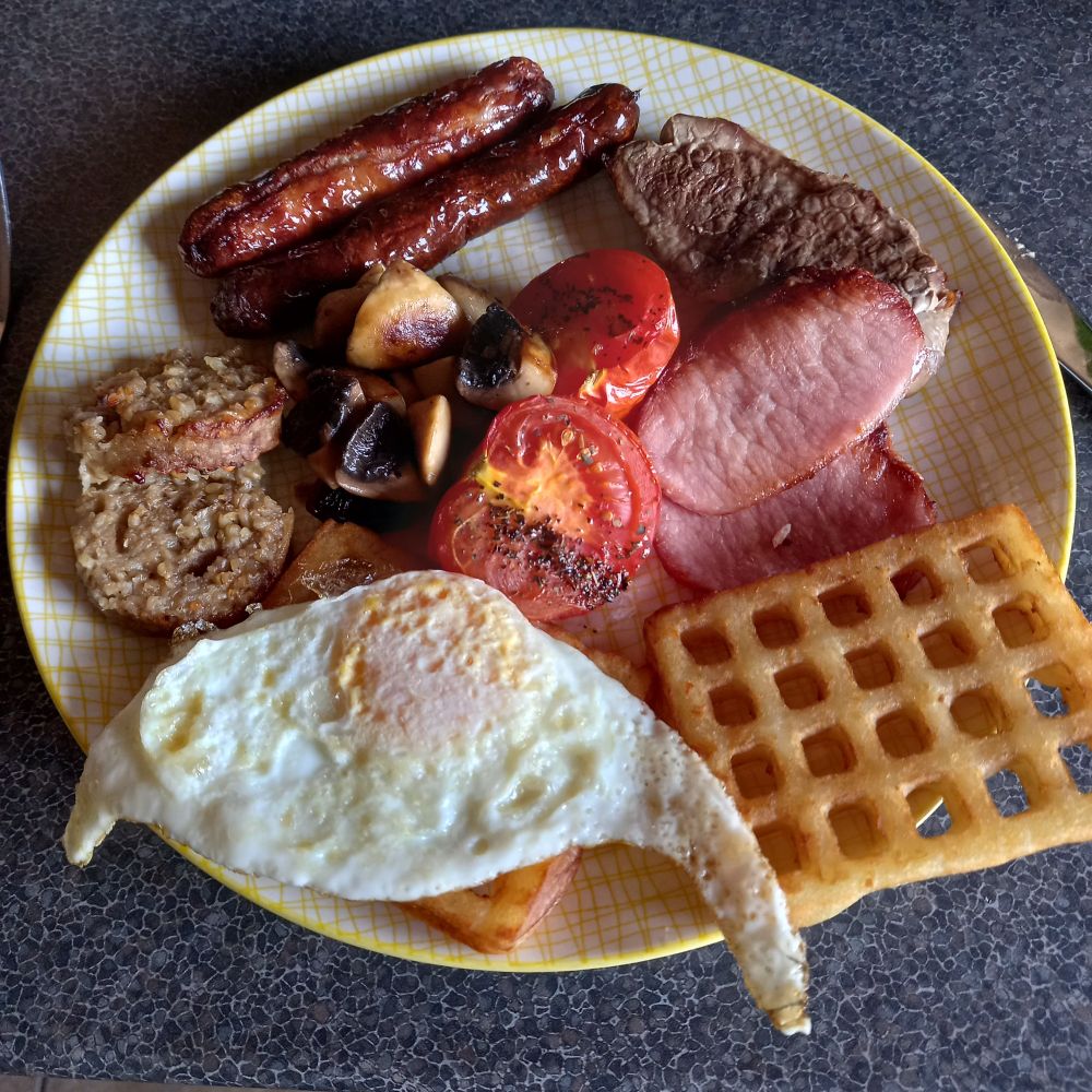 Yellow plate featuring sausages, rashers, pudding, lamb chop, mushrooms, tomatoes, potato waffles and a fried egg. 