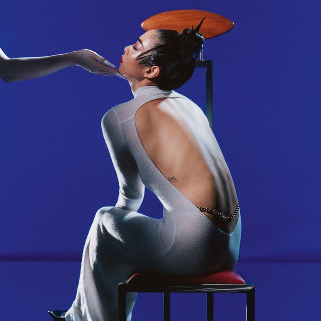 Rina Sawayama’s album cover for Hold the Girl. She sits on a chair in a white dress in front of a blue background. A hand out of frame holds up her chin.