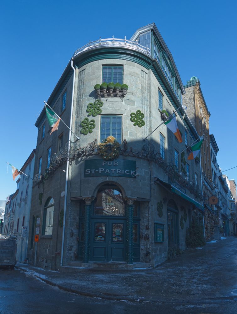 Photo of an Irish Pub, “Pub St-Patrick,” in Quebec City. The 3-story building is on a corner and is triangular in shape with green double doors to the front; 2 green, white and orange flags of Ireland flying from the 2nd storey.