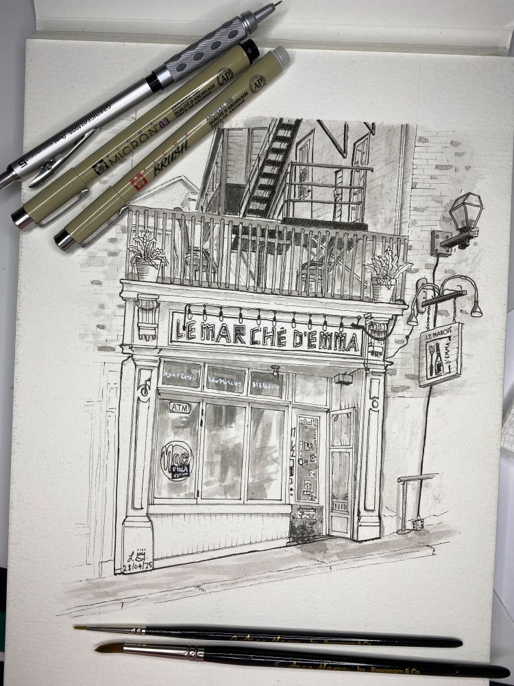 Black and white ink sketch of a small grocery store in between two buildings with a balcony on top. There are pens, a mechanical pencil and paint brushes on top of the sketch.