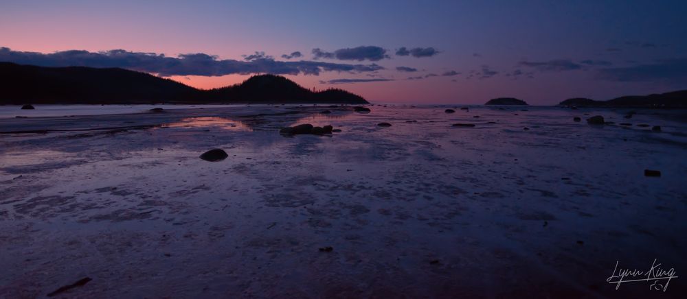 A sunset photo with a purplish blue and orange pinkish sky reflected at low tide in the St. Lawrence Seaway. There are also silhouetted mountains cropping up in the horizon and sporadically placed boulders on the sea floor.