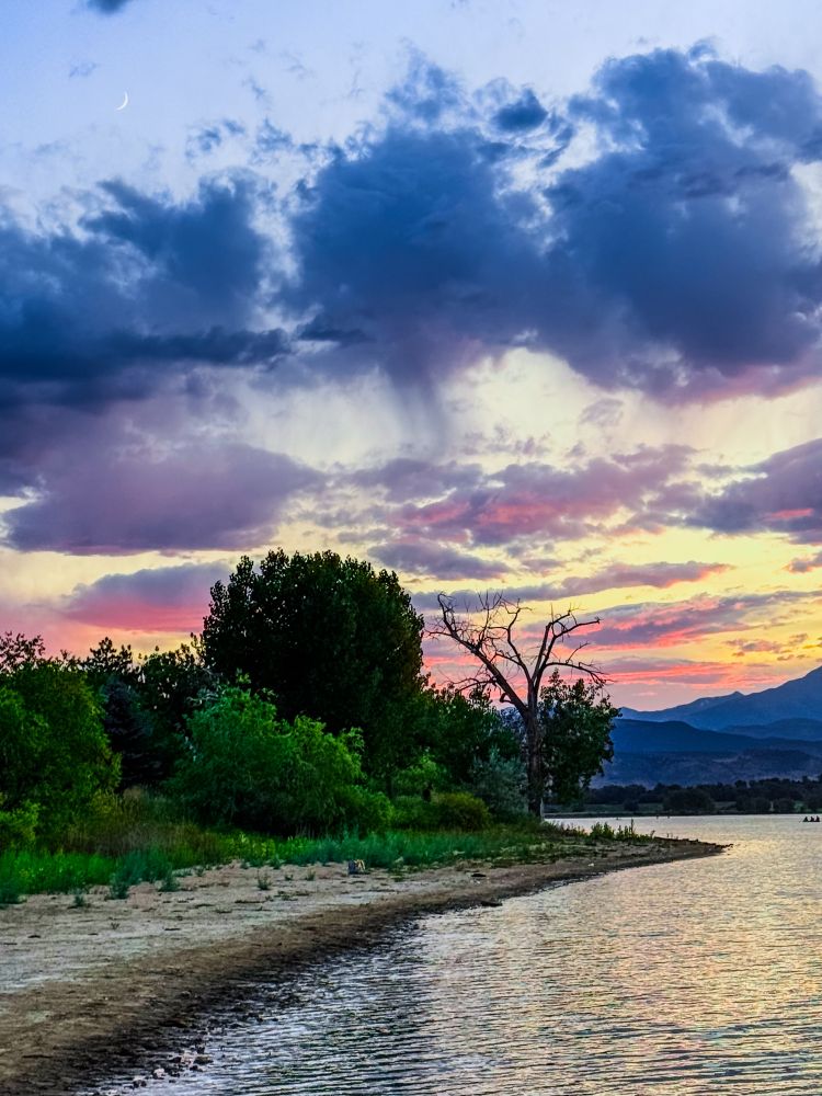 Looking out over a narrow spit of land stretching into a tranquil lake, framed by distant mountains and a sky full of contrast. Storm clouds gather, their deep grays swirling with drama, while a radiant sunset paints strokes of orange, rose, and gold across the horizon. Above it all, a delicate crescent moon rises in the fading blue sky—subtle yet powerful, symbolizing renewal and the quiet strength of beginnings. The land, water, and sky mirror life’s complexity: calm and chaos, darkness and beauty, all held in one frame. A visual meditation on strength, healing, and the layered truth of being alive. Captured in the natural beauty of Colorado.


photography, nature, outdoors, moon, crescent moon, mountains, lakes, Colorado, Colorado photography, healing, wellness, well-being, spirit, spirituality, stormy sky, sunset, layered sky, landscape photography, inspirational quote, resilience, strength, wholeness, nature healing, health, sunset photography, storm clouds, emotional healing, symbolism, water, contrast, reflection, personal growth, mindful living, serenity, moonrise, poetic moments, Instagram poetry, photo art, peace, grounded beauty, earth medicine, inspiration, mountains, ocean by Eric Meleney @redcranehealth