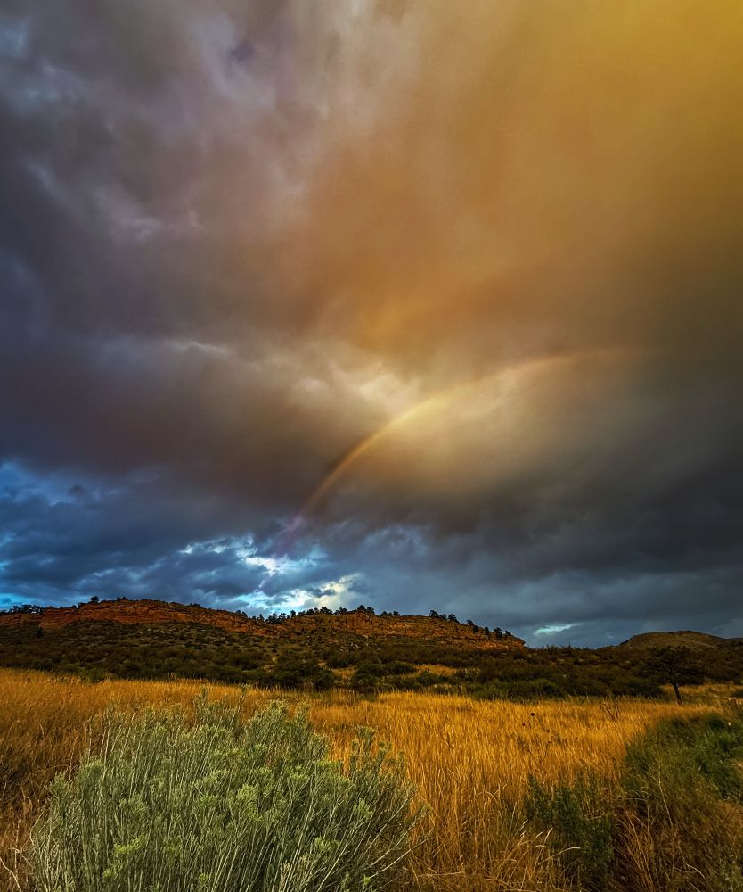Stormy desert sunset with dramatic clouds parting as a rainbow glows through, framed by rocky ground, dry golden grasses, and green foliage. The scene evokes resilience, flow, and growth, a reminder of healing and transformation emerging through change. Keywords: stormy sky, rainbow, sunset clouds, Colorado, nature photography, spiritual journey, wellness.