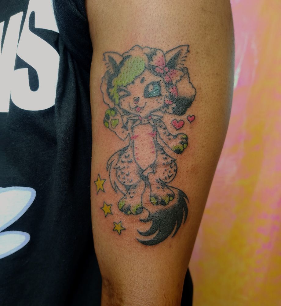 A tattoo on the bicep of a fursona based on a Creeper. He has a bow on his ear, green bangs and a pink raccoon tail in his hair, as well as spot markings and top surgery scars. Surrounded by hearts and stars, with a fluffy tail flowing downwards. He looks cheerful, waving.