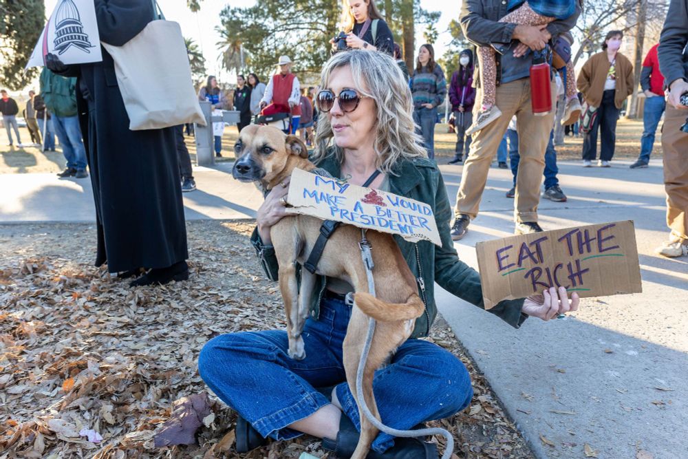 A woman with long blond hair, wearing jeans and green jacket is holding her dog in her lap as she sits on the ground during a rally/march in Tucson, AZ on Inauguration Day, 01/20/25. Her dog has a sign on its back that says "My (poop emoji) would make a better president." The woman is holding a sign that says "Eat the rich." Image by the author of this post, Kathleen Dreier.