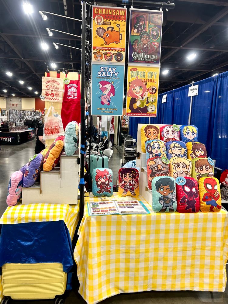 A side view photograph of a convention table set up. Plush pillows, notebooks, charms, and other merchandise are displayed on a table with a yellow checkered tablecloth. There is also an open catalog of pillow designs on the table. Posters are displayed overhead on a photostand display.