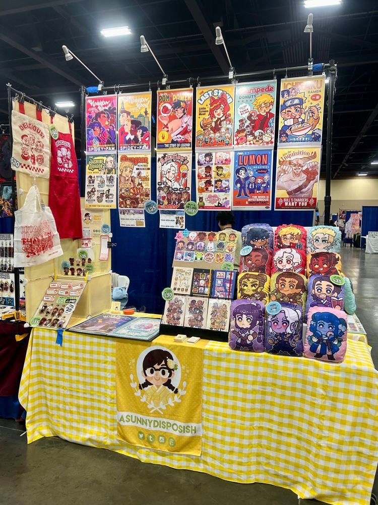 A photograph of a convention table set up. Plush pillows, notebooks, charms, and other merchandise are displayed on a table with a yellow checkered tablecloth. Posters are displayed overhead on a photostand display. The table banner reads “A Sunny Disposish”