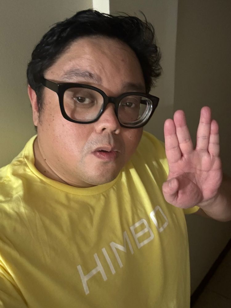 A fat* Brown male with thick black hair, eyeglasses with a thick black frame. He is raising his hand to make the gesture known as the Vulcan greeting (“live long and prosper”). He is making an unserious expression on his face, a mixture of feigned sexiness and genuine confusion, as he pouts his thin and nearly nonexistent lips. He wears a yellow ring-collar t-shirt that says the word “HIMBO” in white lettering.