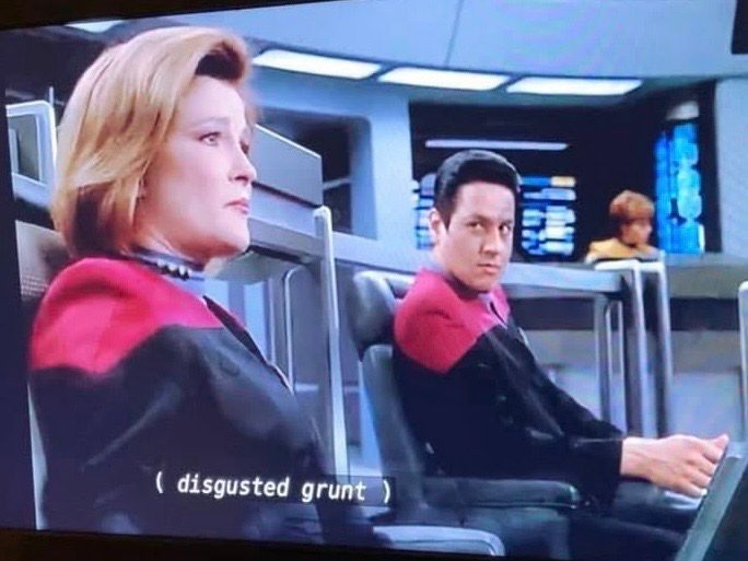 Three people are pictured. From L-R: Captain Janeway, whose facial expression is in mid-reaction; Commander Chakotay, who is staring expectantly at Captain Janeway; and an unknown crewman in the background. A closed captioning display says, ( disgusted grunt ).