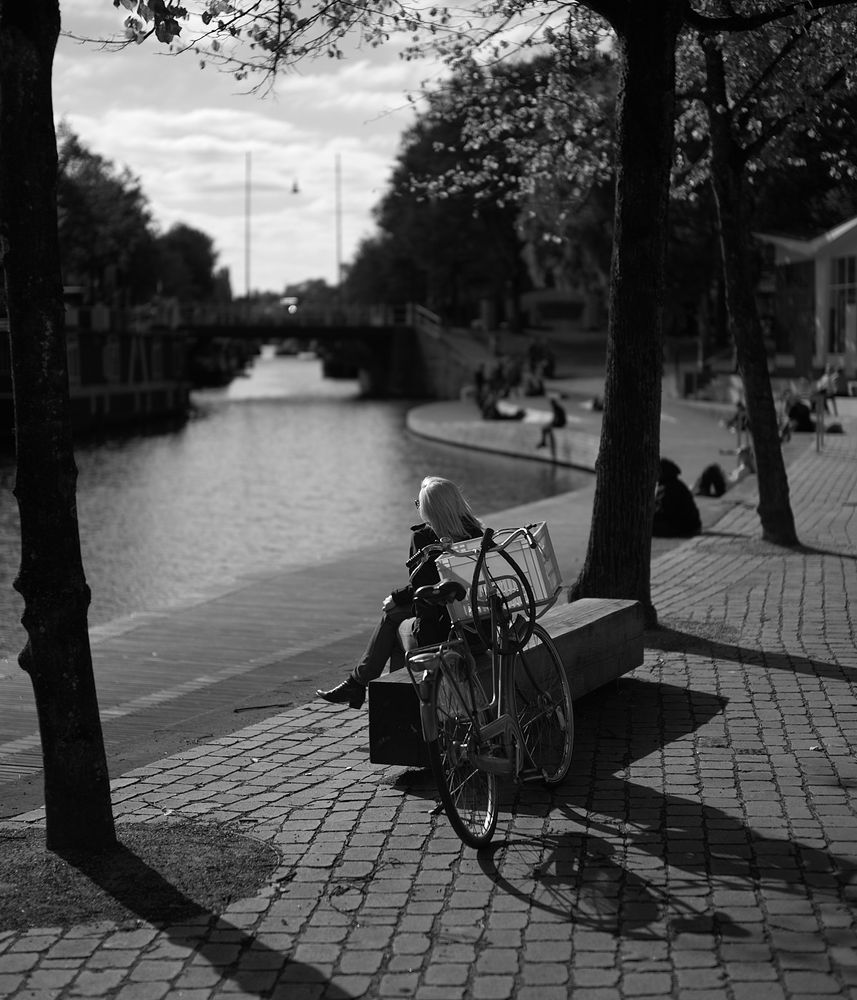 A woman sits on a bench by a canal, looking out over the water. A bicycle with a basket rests nearby. The scene is in black and white, featuring trees and people in the background along the water's edge.