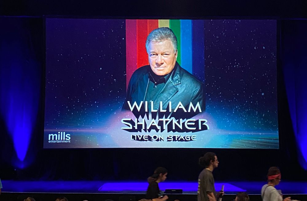 A large projection screen on a theater stage with a photo of William Shatner and the caption “William Shatner live on stage”