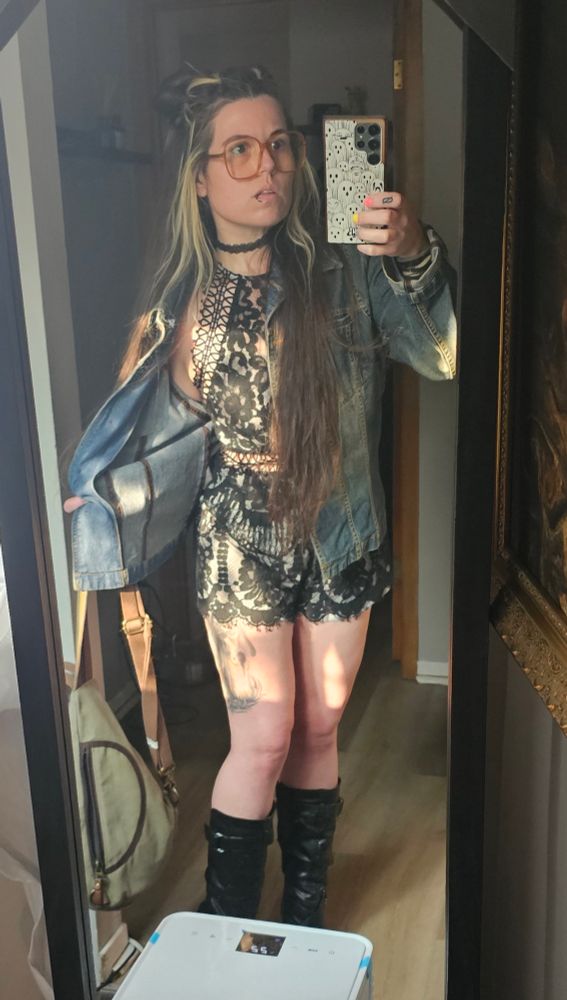Doxy posing in front of the mirror, showcasing a lacy black romper under a denim jacket with mid-calf, black, heeled boots