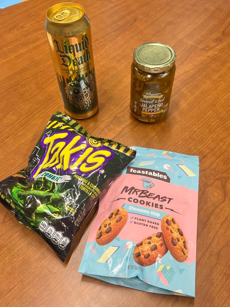 A picture of a bag of Zombie flavored Takis, Mr. Beast chocolate chip cookies, a Liquid Death brand tea and a jar of jalapeño peppers 