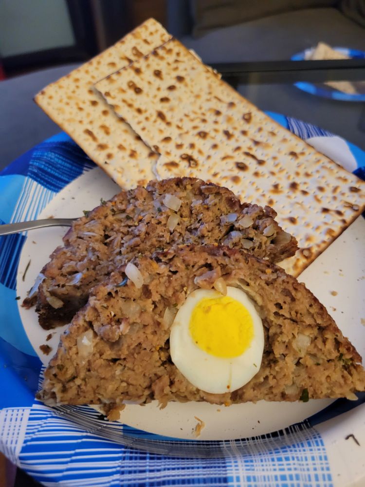 Two slices of meatloaf, called klops, sit on a blue & white paper plate with a broken board of matzah - unleavened bread typically eaten during Passover. One slice has a hard-boiled egg slice visible in it.