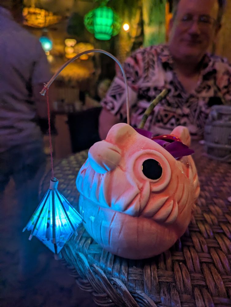 A pink angler fish tiki mug with a blue glowing tiki cocktail lantern decoration dangling in front of it enticingly