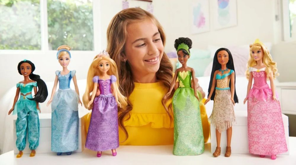A child smiling behind 6 Disney Princess dolls stood on a table.