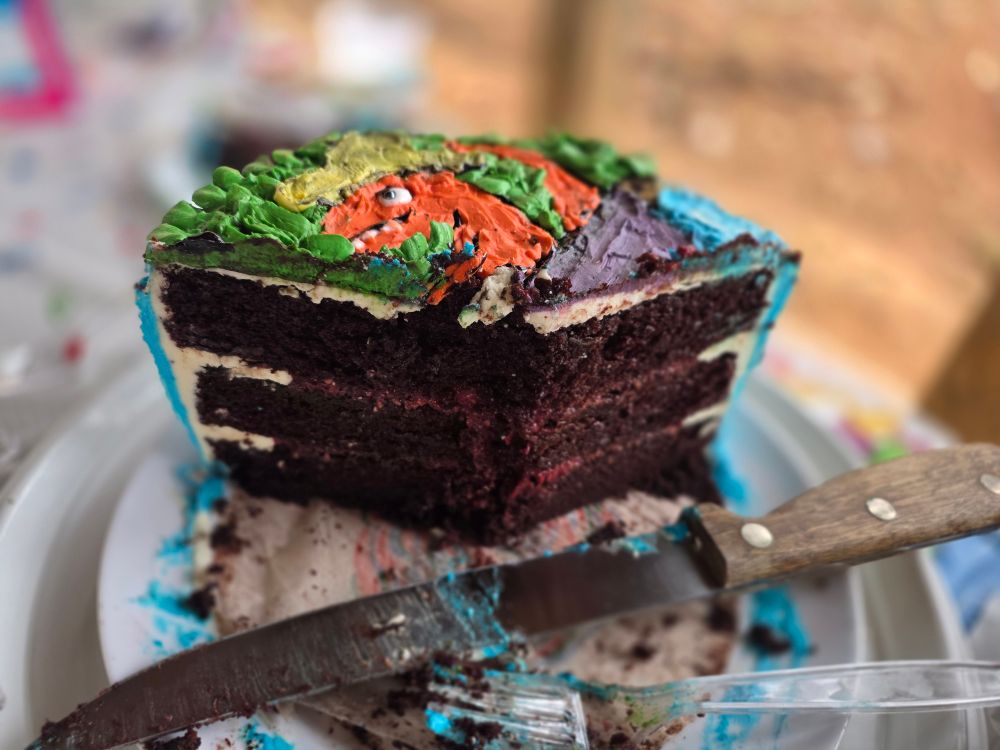 Photo of a third of a 3-layer chocolate cake that has a frosting drawing of an orange t-rex in a yellow hat on it.