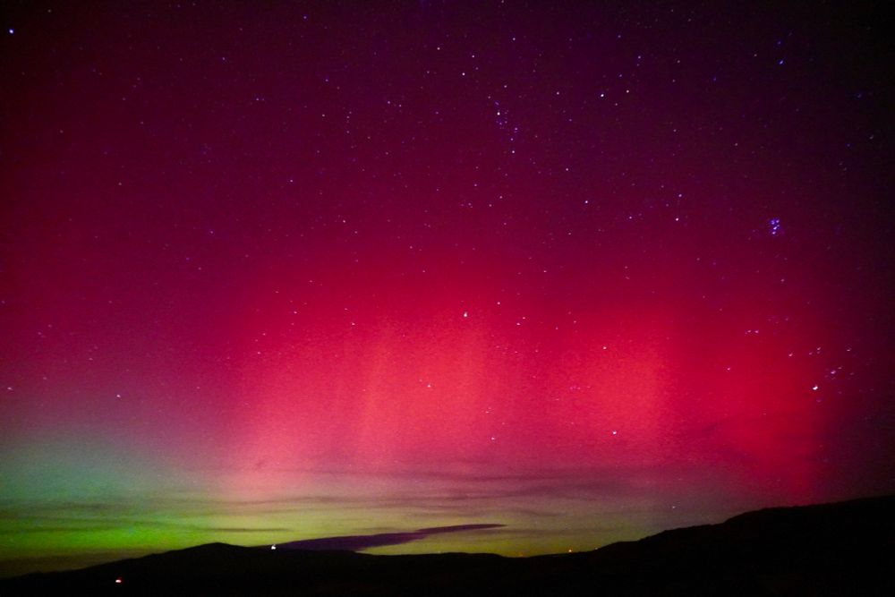 A dark silhouette of hills rests beneath a star-filled night sky. Above the hills, an aurora displays a band of green light that blends upward into a vibrant reddish-pink and magenta glow, ultimately dissolving into the dark purple cosmos.