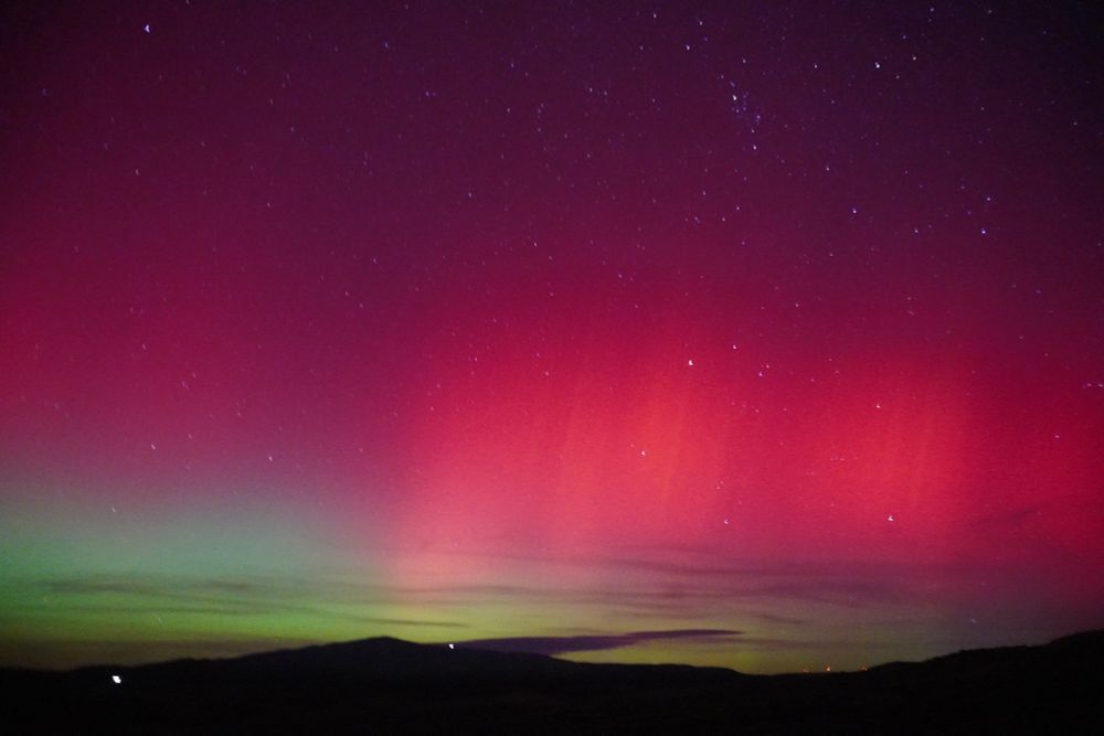 A night sky filled with stars features a colorful aurora. A dark silhouette of hills rests at the bottom, above which a band of green light transitions into a brilliant, deep reddish-pink and magenta glow that fades into the dark purple cosmos above.