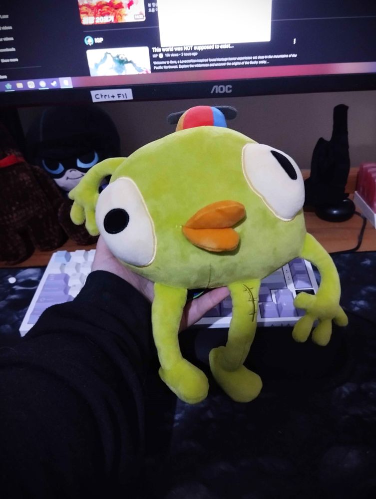 Small Makeship plushie, about two small hand sizes of Bing Bong from the game Peak. He is green with two large eyes and an orange mouth, wearing a red, blue and yellow propeller hat.