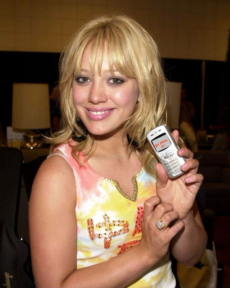 hilary duff from years ago wearing a yellow tie dye tank top and holding up an old cellphone with the text “go piss, girl” on it 
