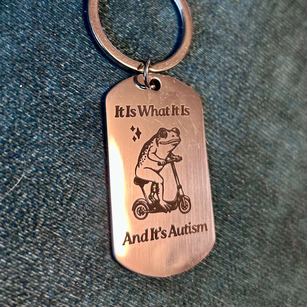 A steel keychain with a carving of a frog on a scooter. Text reads It Is What It Is. And It's Autism.