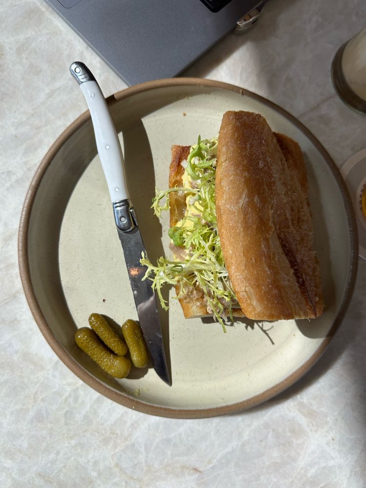 It top down shot of a sandwich, a sharp knife, and three corners sitting on the ceramic plate on a marble counter next to a MacBook.