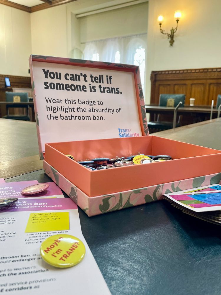 A box full of badges in a room in Parliament which say Maybe I'm Trans? in a box which says You can't tell if someone is trans. Wear this badge to highlight the absurdity of the bathroom ban. 
