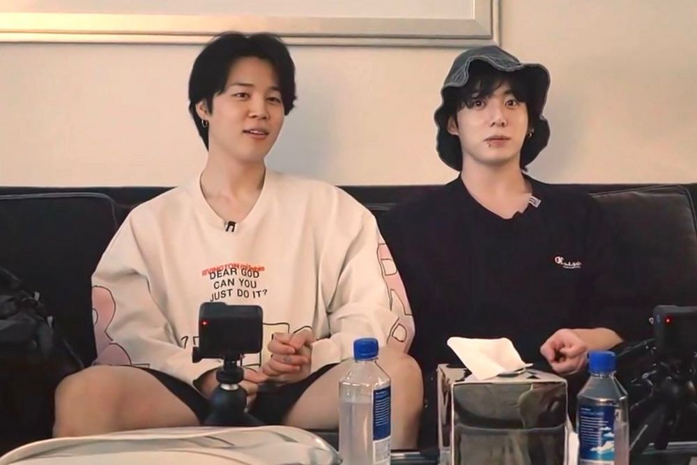 Jimin and Jungkook from the first episode of are you sure. They are in the hotel room, Jimin wearing a white long sleeve tee and Jungkook wearing an oversized black tee and his bucket hat. They are holding hands for some unknown reason
