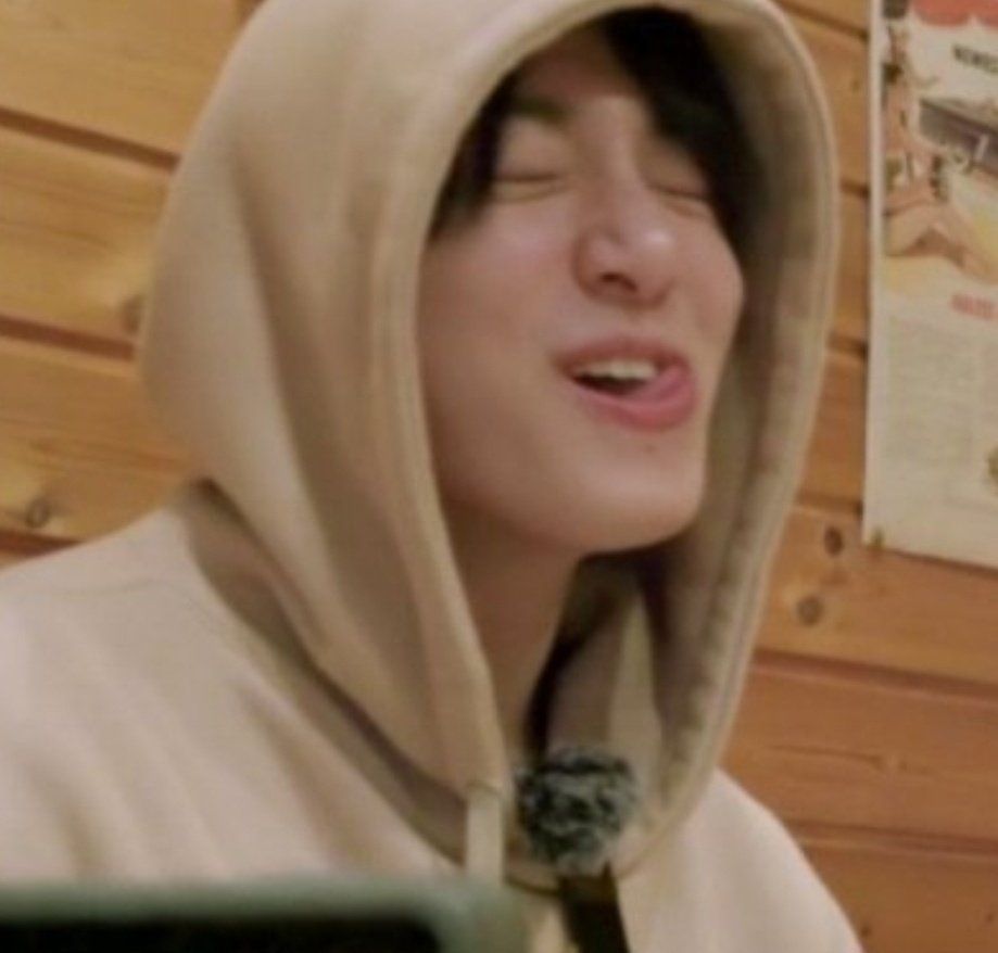 Jungkook wearing a cream hoodie, sitting in a restaurant with Jimin during ays sapporro. He's laughing with his eyes closed, nose scrunched and his tongue is poking out one corner of his mouth.