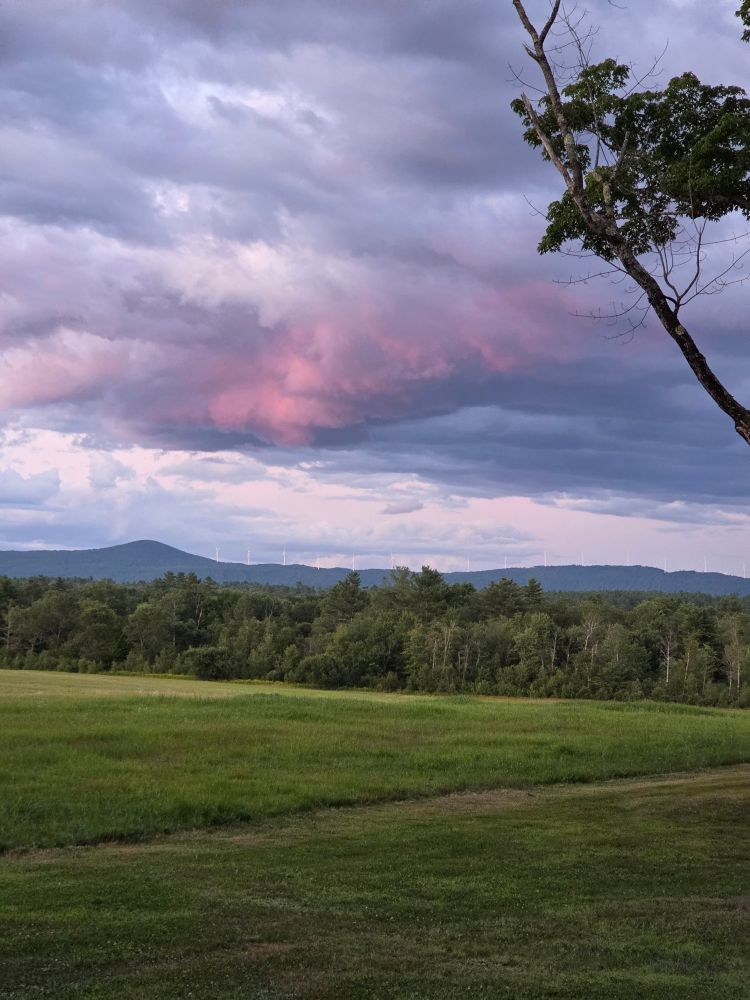 Colorful clouds over a green field with trees and mountains beyond. 