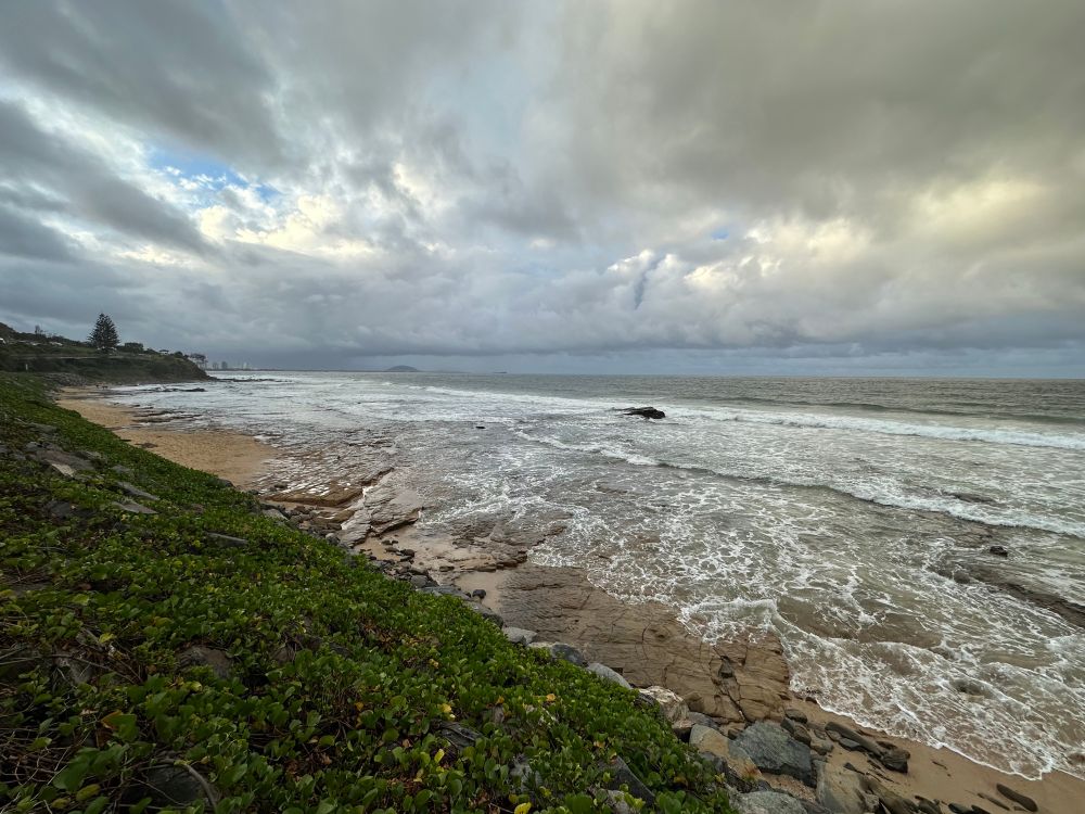 Stormy skies over a rocky beach. A grassy hill leads to a headland 