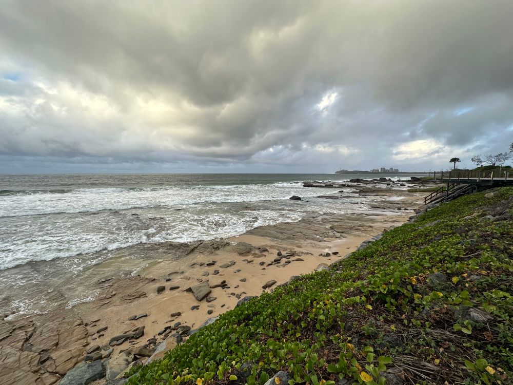 Stormy skies over a rocky beach. A few trees and stairs plus a headland with buildings in the far distance