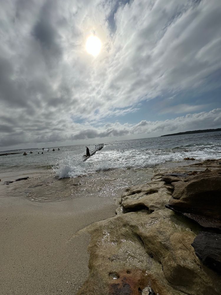 Over cast sky with sandy and rocky beach as small waves crash over man made rock pool